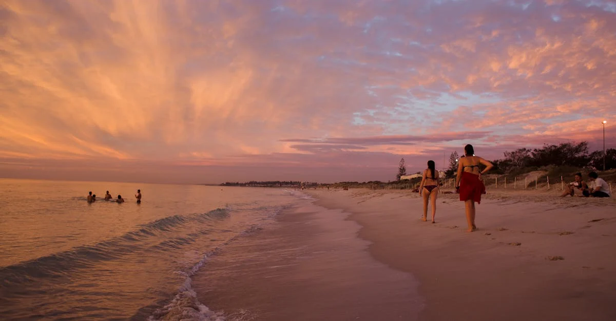 Two women walking along a serene beach at sunset, vibrant sky reflections.